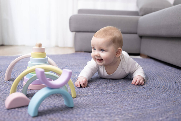 Blanke pasgeborene van 3 maanden oud die probeert te kruipen. Blij kijkt naar prachtige houten multi-gekleurde speelgoed. De baby ligt op zijn buik op een blauw rieten tapijt naast de bank. Kinderontwikkeling, warm vloerconcept. - Foto, afbeelding