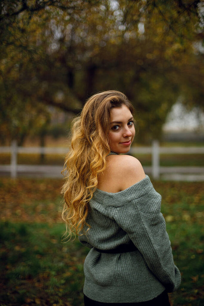 the girl playfully looks around the outside. young woman in a warm jacket in the park or garden. cheerful girl stands in the countryside in cloudy weather - Photo, Image