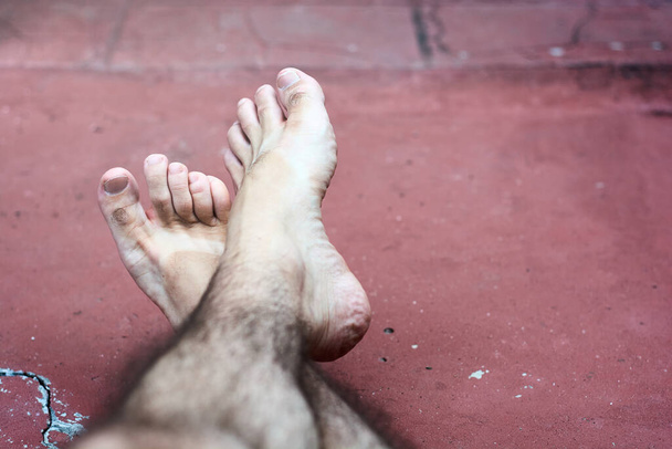 Close up of crossed bare feet of caucasian young man lying on red ground. Concept of relaxation and feet care. - Photo, Image