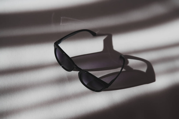 Κομψά μαύρα γυαλιά ηλίου σε λευκό φόντο με σκιές. Πλαστικά ποτήρια με κλίση σε μια ηλιόλουστη μέρα. Άποψη από ψηλά. - Φωτογραφία, εικόνα