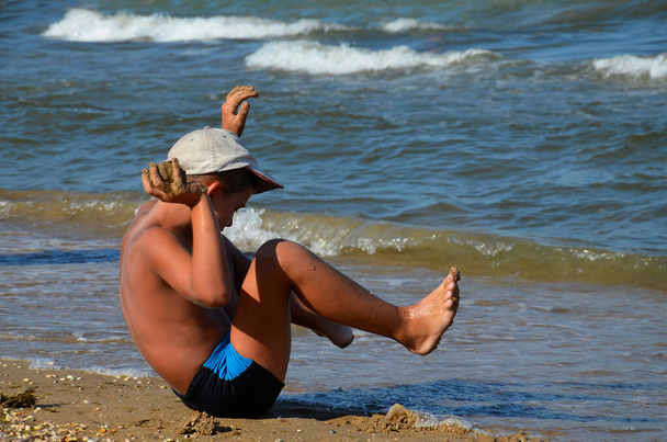 Счастливый мальчик играет на песчаном пляже на фоне моря. Счастливый мальчик сидит на морском пляже. Вид сбоку. Саммер. Семейный отдых. Путешествие. - Фото, изображение