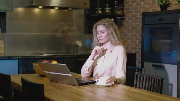Girl sits thoughtfully in kitchen in evening drinking coffee and looking laptop screen. woman is engaged in successful business development remotely. freelancer edits text of message - Footage, Video