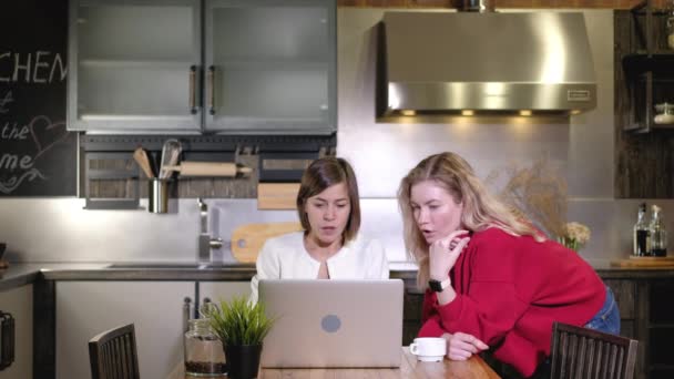 Dos chicas caucásicas trabajan juntas en casa remotamente. Las mujeres de negocios discuten proyecto de trabajo en la computadora en la mesa de la cocina. Intercambio de ideas y lluvia de ideas. Debate empresarial y toma de decisiones - Metraje, vídeo