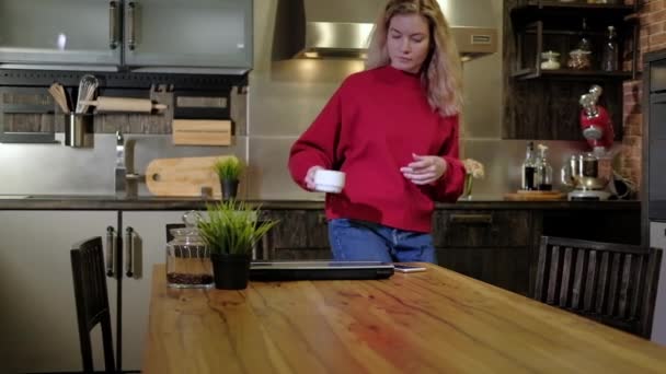 Freelancer girl starts working in kitchen at home in morning. woman with mug of hot, invigorating coffee comes to table with a laptop, opens it and starts working at the workplace. Remote work at home - Footage, Video