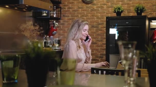 Συγκεντρωμένη νεαρή κοπέλα επικοινωνεί smartphone με τον πελάτη το βράδυ στην κουζίνα στο διαμέρισμα και πληκτρολογήστε μήνυμα στο chat στο laptop. Γραφείο στο διαμέρισμα. γυναίκα ελεύθερος επαγγελματίας μιλάει στο τηλέφωνο εξ αποστάσεως - Πλάνα, βίντεο