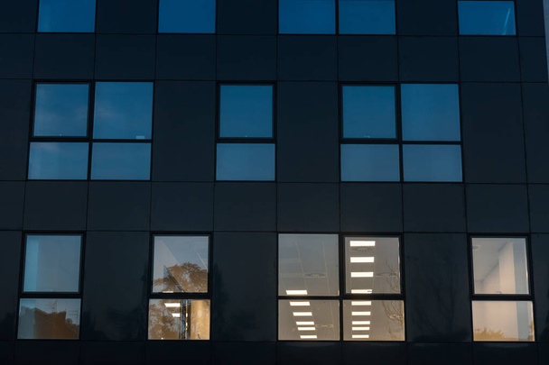 Fachada de um edifício de distrito de negócios, um edifício alto, à noite, à noite, com apenas algumas janelas acesas, para funcionários que trabalham até tarde neste edifício de escritórios - Foto, Imagem