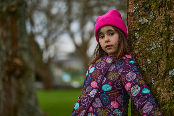 Menina caucasiana de pé perto de uma árvore. Com roupas casuais e um chapéu rosa. Em um parque natural. - Foto, Imagem