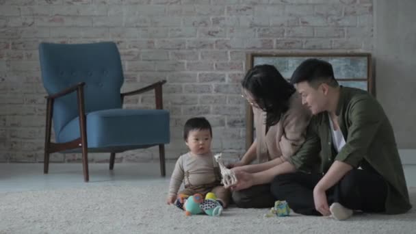 Genç Asyalı aile. Bir anne ve baba oturma odasında yerde yeni doğmuş küçük oğullarıyla oynuyorlar. Sevgi dolu ebeveynler çocuklarına oyuncak verir ve mutlu bir şekilde oyunu izlerler.. - Video, Çekim