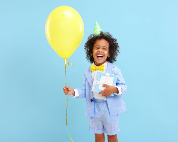 Cheerful little black boy with curly hair in stylish suit and party hat smiling while standing against blue background  with yellow balloon and present in hands - Photo, Image