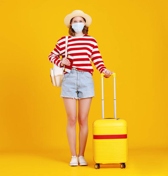 Cuerpo completo de joven viajera irreconocible en ropa casual y mascarilla de pie contra amarillo con maleta y mirando a la cámara - Foto, Imagen