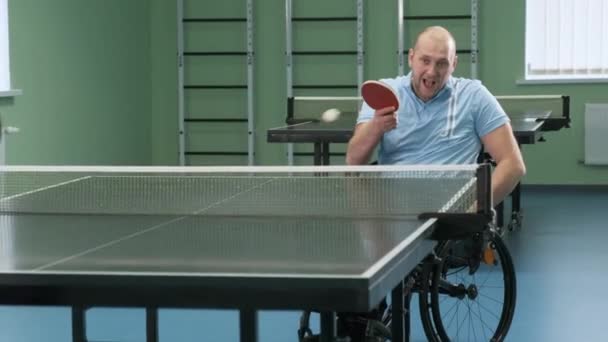Egy tolószékes férfi pingpongozik. Fogyatékos emberek asztaliteniszeznek. A fogyatékkal élők rehabilitációja. Paralimpiai sport. - Felvétel, videó