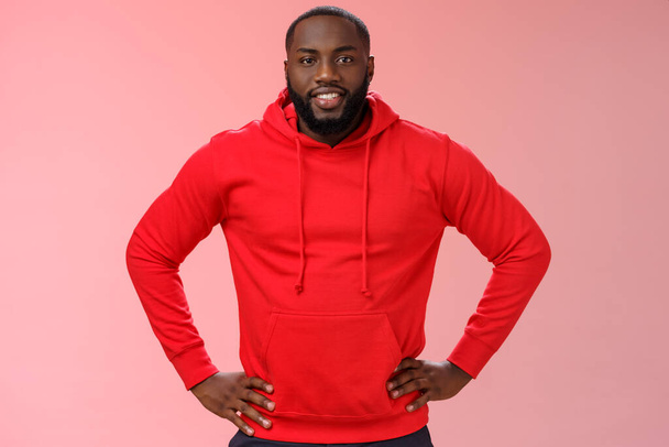 Nuttige aantrekkelijke vriendelijke bebaarde Afrikaans-Amerikaanse vriend dragen rode hoodie hold handen taille glimlachen wilt helpen vragen wat te doen, helpende hand, staande zelfverzekerde assertieve roze achtergrond - Foto, afbeelding