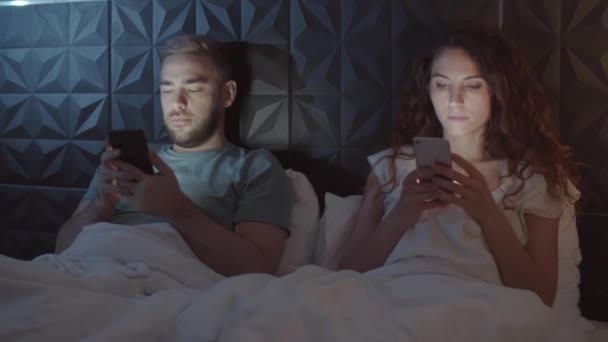 Высокая дуга снимка молодого кавказского мужа и жены в пижамах, сидящих молча в постели ночью, опирающихся на спинку кровати, держащих смартфоны и выглядящих полностью погруженными - Кадры, видео