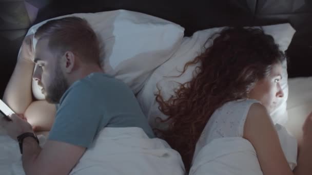 Pas-up záběr rozčarovaný mladý běloch manžel a manželka ležící v posteli spolu v noci, zády k sobě, plně zapojeni do používání svých chytrých telefonů - Záběry, video