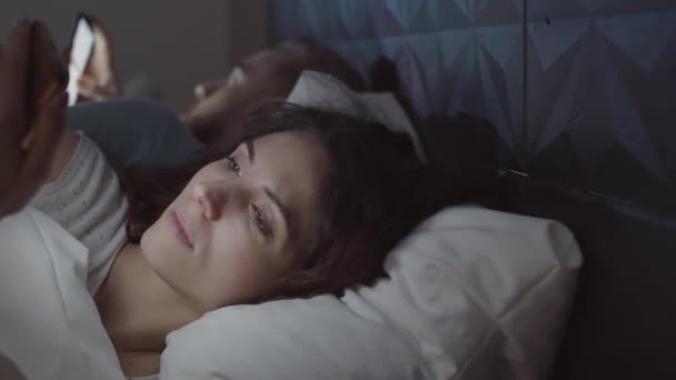 Chest-up panning záběr krásné mladé ženy ležící v posteli v noci s manželem, tvář osvětlena chytrým telefonem, dívá se přes sociální sítě a usmívá - Záběry, video