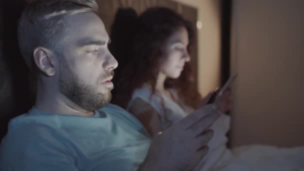 Rinta-up laukaus nuori valkoihoinen mies makaa sängyssä, pitämällä älypuhelin käsissä, katsomalla näytön ja kirjoittamalla viestin sosiaalinen verkosto, ja hajamielinen vaimo istuu vieressä - Materiaali, video