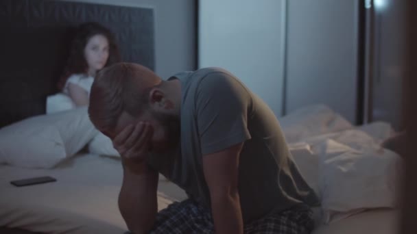 Medium shot van uitgeputte jonge blanke man zitten op de zijkant van bed, met smartphone naast kussen, en wrijven voorhoofd, en boze vrouw staren naar zijn rug - Video