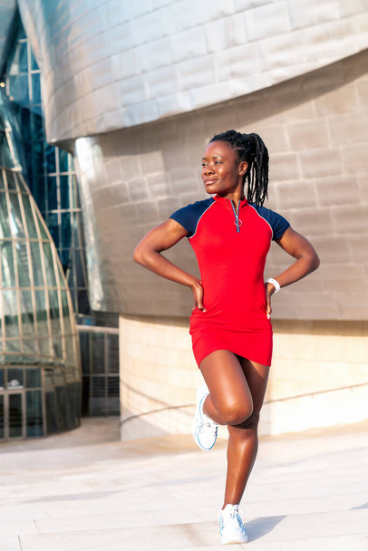 černá afro dívka s úsměvem pózuje na ulici moderního města v barevných červených šatech - Fotografie, Obrázek