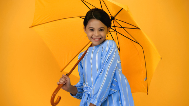χαρούμενο παιδί με μπλε φόρεμα που στέκεται κάτω από ομπρέλα απομονωμένο σε κίτρινο - Φωτογραφία, εικόνα