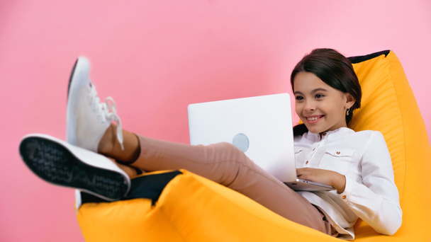 bambino sorridente che usa il computer portatile mentre riposa sulla sedia del sacchetto del fagiolo isolato su rosa - Foto, immagini
