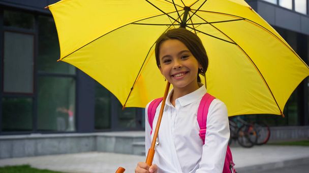 colegiala feliz con paraguas de pie cerca del edificio - Foto, Imagen