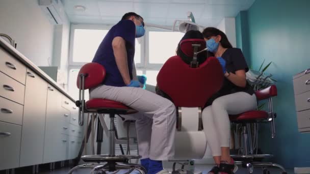 Οι γιατροί οδοντίατροι γυναίκα και ο άνθρωπος με ιατρικές μάσκες εξυπηρετούν τον ασθενή στην οδοντιατρική κλινική - Πλάνα, βίντεο