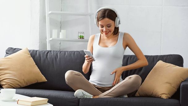 boldog terhes nő ül a kanapén, zenét hallgatni és beszélgetni okostelefonon - Fotó, kép