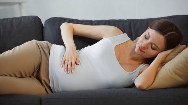 красивая беременная женщина, прикасающаяся к животу, отдыхая дома на диване - Фото, изображение