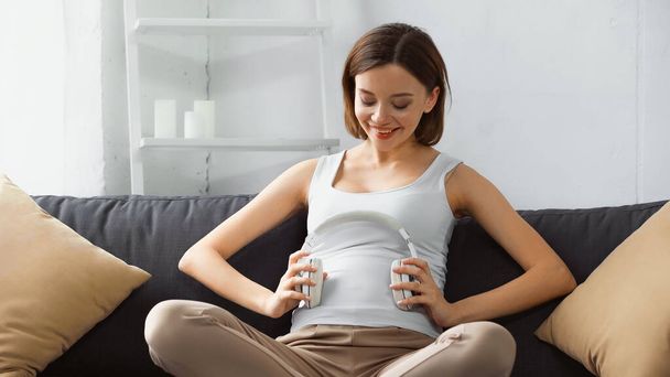 glücklich schwangere Frau hält Kopfhörer in Bauchnähe, während sie auf der Couch sitzt - Foto, Bild