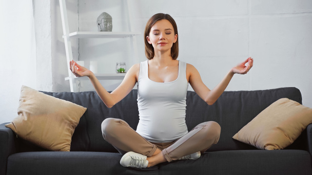 junge schwangere Frau meditiert mit geschlossenen Augen auf dem heimischen Sofa - Foto, Bild