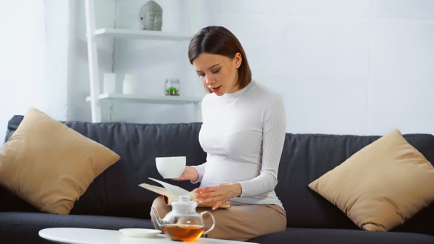 pregnant woman reading book and drinking tea on sofa at home - Photo, Image