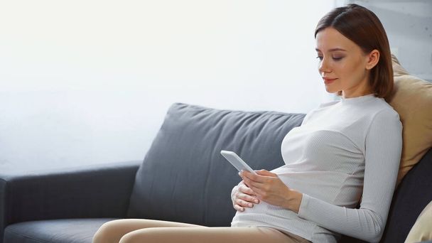 молодая беременная женщина сидит на диване и отправляет сообщения на смартфон - Фото, изображение