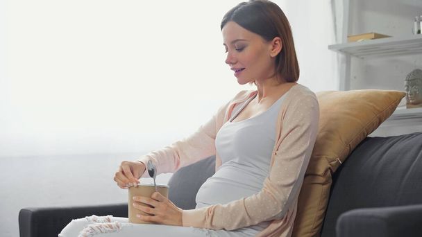 junge schwangere Frau öffnet Eis, während sie auf dem Sofa sitzt - Foto, Bild