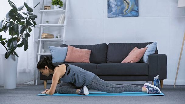 mujer joven flexible en ropa deportiva que se extiende en la alfombra de fitness en casa  - Foto, Imagen