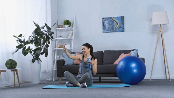 heureux jeune femme en vêtements de sport assis sur tapis de fitness et de prendre selfie à la maison  - Photo, image
