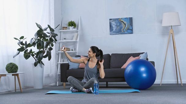 veselá mladá žena ve sportovním oblečení sedí na fitness podložce a brát selfie doma  - Fotografie, Obrázek