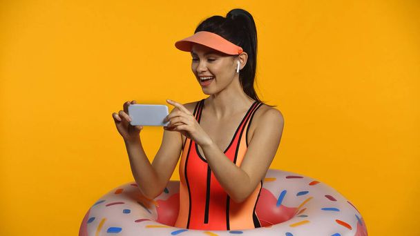 Vidám fiatal nő vezeték nélküli fülhallgató fürdőruha és felfújható gyűrű okostelefon elszigetelt sárga - Fotó, kép