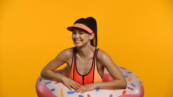 Cheerful young woman in swimsuit and inflatable ring smiling isolated on yellow - 写真・画像