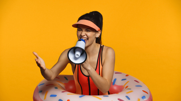 Joyeux jeune femme en maillot de bain et anneau gonflable criant en mégaphone isolé sur jaune - Photo, image