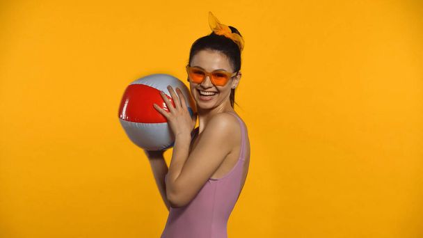 Ευτυχισμένη νεαρή γυναίκα με μαγιό κρατώντας φουσκωτή μπάλα απομονωμένη στο κίτρινο - Φωτογραφία, εικόνα