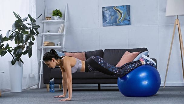 Spor kıyafetli genç esmer kadın evde fitness topuyla egzersiz yapıyor.  - Fotoğraf, Görsel
