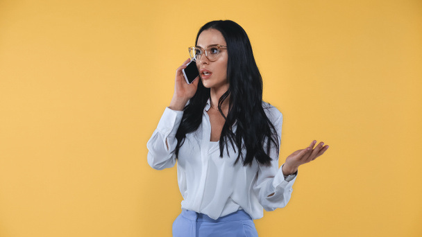обескураженная деловая женщина жестикулирует, разговаривая по мобильному телефону изолированы на желтый - Фото, изображение