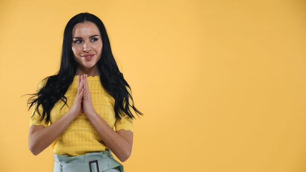 positive brunette woman standing with praying hands isolated on yellow - Photo, Image