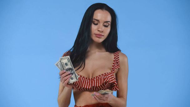 mooi, brunette vrouw in badpak tellen dollar bankbiljetten geïsoleerd op blauw - Foto, afbeelding