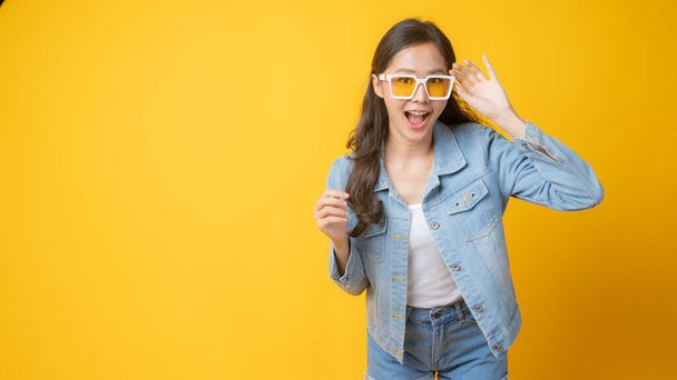 young cute Asian woman wearing yellow fashion glasses in jeans posing and smiled, Thai joyful and beautiful girl isolated on yellow background - Photo, Image