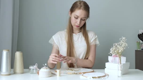 Ein junges Mädchen mit langen Haaren sitzt auf dem Arbeitsplatz in der hellen Werkstatt und stickt mit Perlen. Die Designerin stellt handgemachten Schmuck her. Hobby Konzept. Selbstständige Freiberuflerin macht Kreativität - Filmmaterial, Video