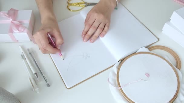 少女は、布に刺繍の絵を作成するためにノートブックにスケッチを描く光のワークショップのワークスペースに座っています。趣味の概念。自己雇用のフリーランスは、創造性を手作り - 映像、動画