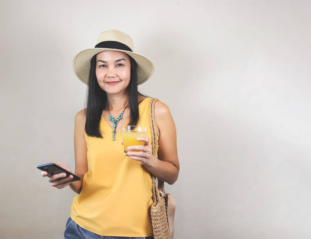 Portret van een gezonde Aziatische vrouw met een geel mouwloos hemd en hoed met mobiele telefoon, een glas geel sap vasthoudend, glimlachend en kijkend naar de camera. Zomer en gezond drankconcept. - Foto, afbeelding