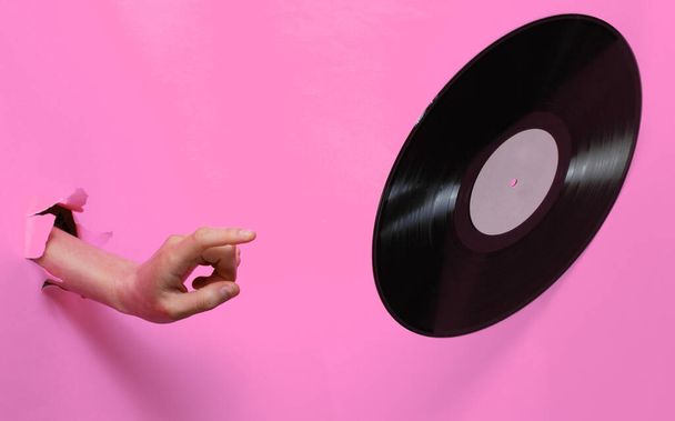 女性の手は、引き裂かれたピンクの紙の背景を通してビニールレコードを回転させます。ミニマルレトロなコンセプト - 写真・画像