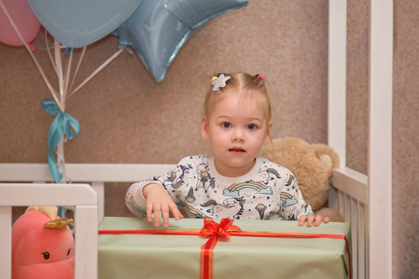 Mała dziewczynka w piżamie otrzymuje prezenty urodzinowe siedząc w białym łóżeczku. Balony w tle - Zdjęcie, obraz